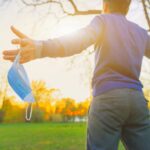 Ihminen ulkona maski kädessä levittää kädet auringon edessä