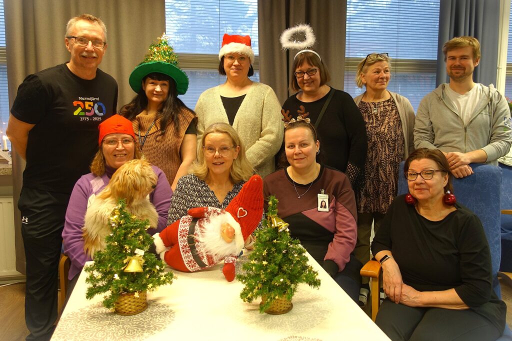 Ryhmä ihmisiä pöydän ääressä, päällään jouluisia asusteita. Edessä pieniä joulukuusia.