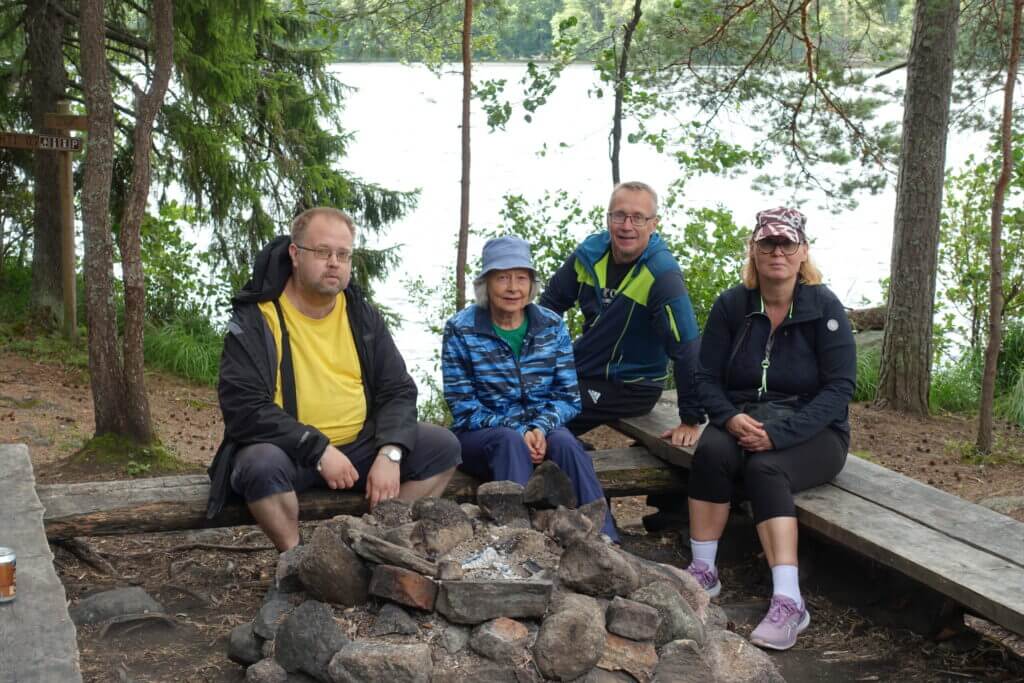 Neljä ihmistä kesäisessä luonnossa nuotiopaikan ympärillä taustalla järvi